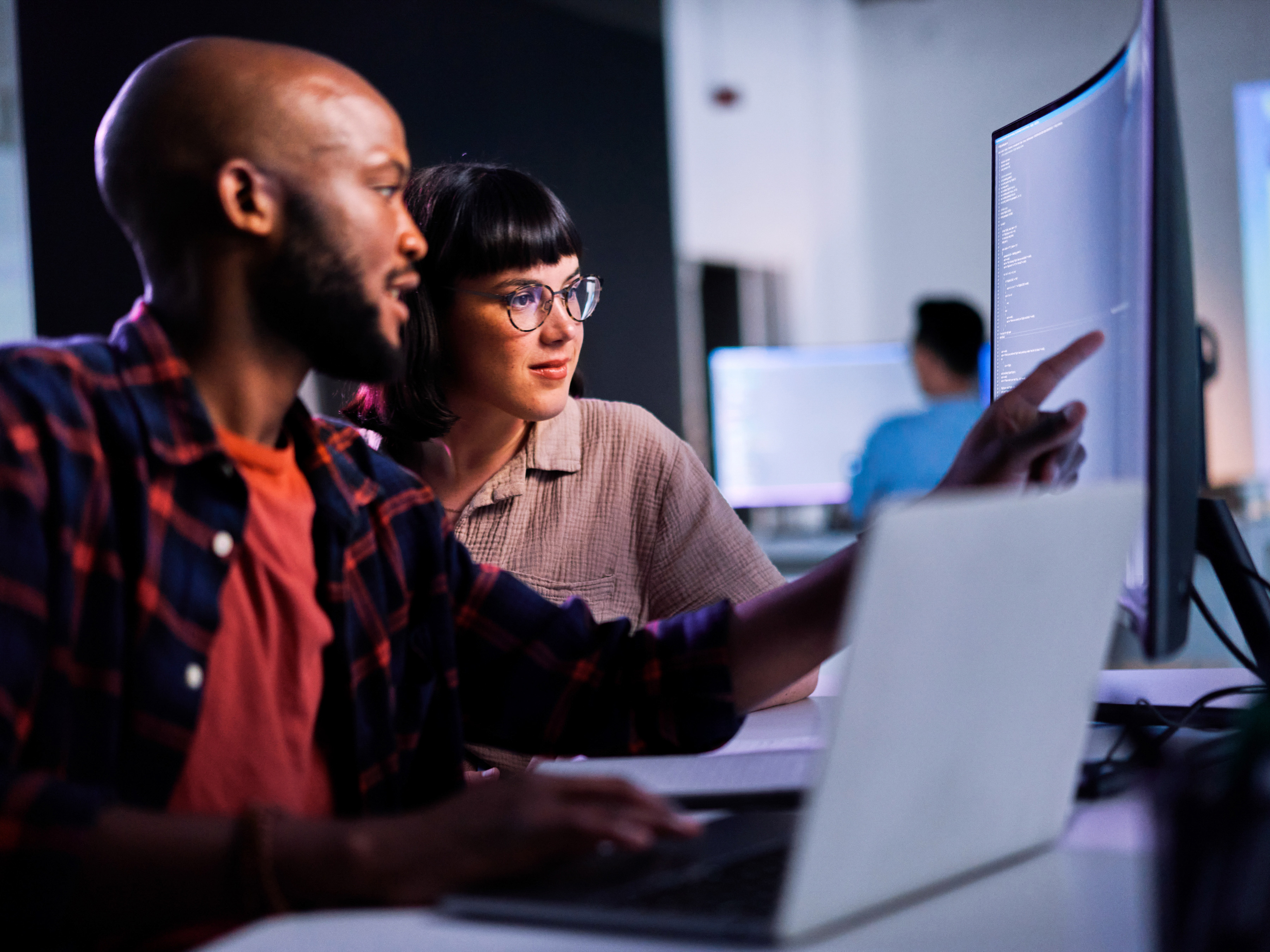 Two employees collaborating in front of a large desktop monitor at work. Two employees collaborating in front of a large desktop monitor at work.
