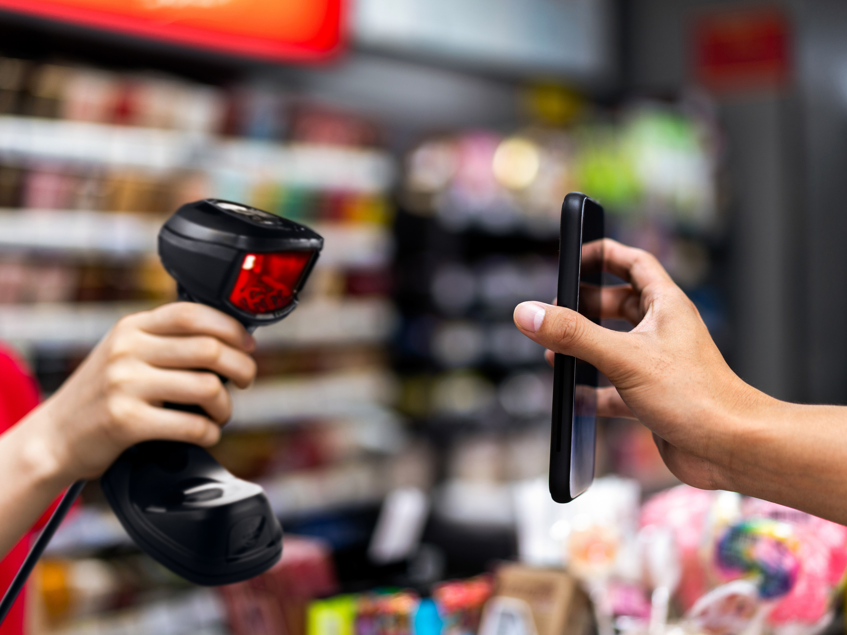 A retail employee using a handheld barcode scanner to scan a mobile application on a shopper's phone during checkout at a store. A retail employee using a handheld barcode scanner to scan a mobile application on a shopper's phone during checkout at a store.