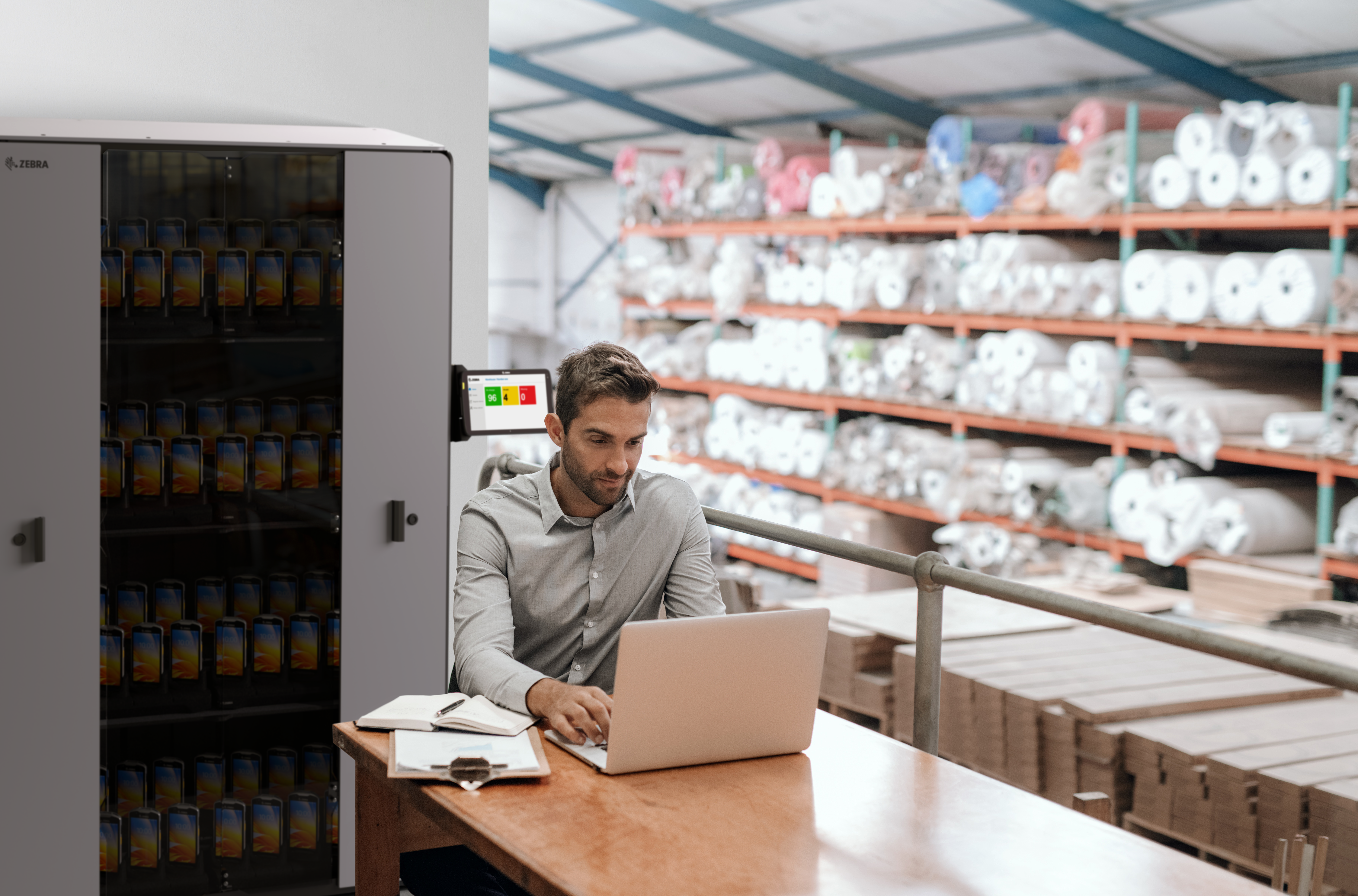 Manager working on a laptop while sitting at his desk in a carpet warehouse with shelves full of stock in the background 