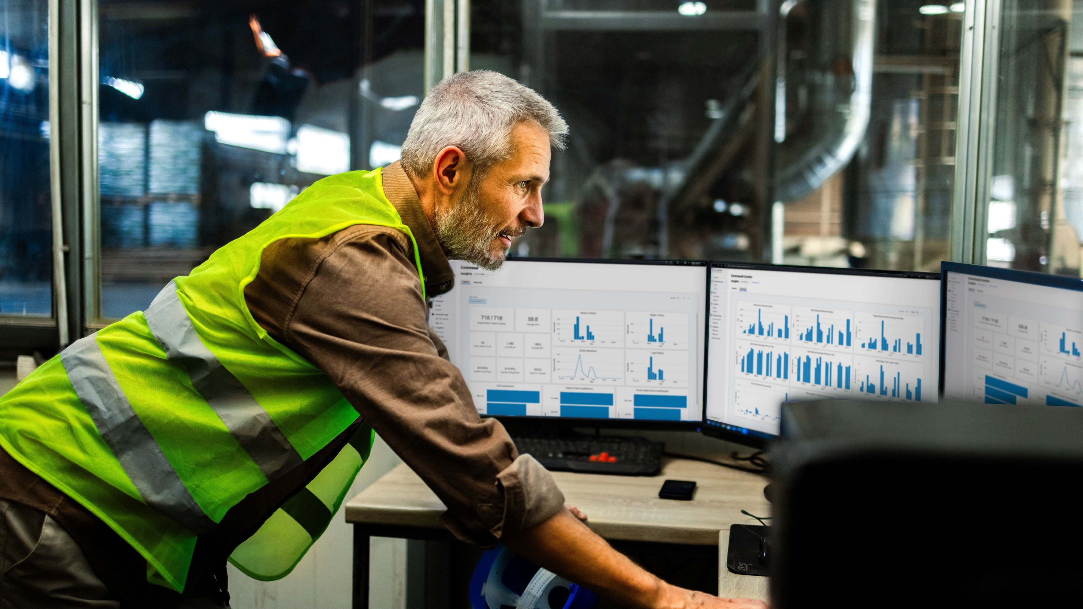 A worker is leaning forward on a desk and examining multiple computer monitors displaying various data and control panels in an industrial office. Image of facility manager reviewing the Symmetry software command center analytics