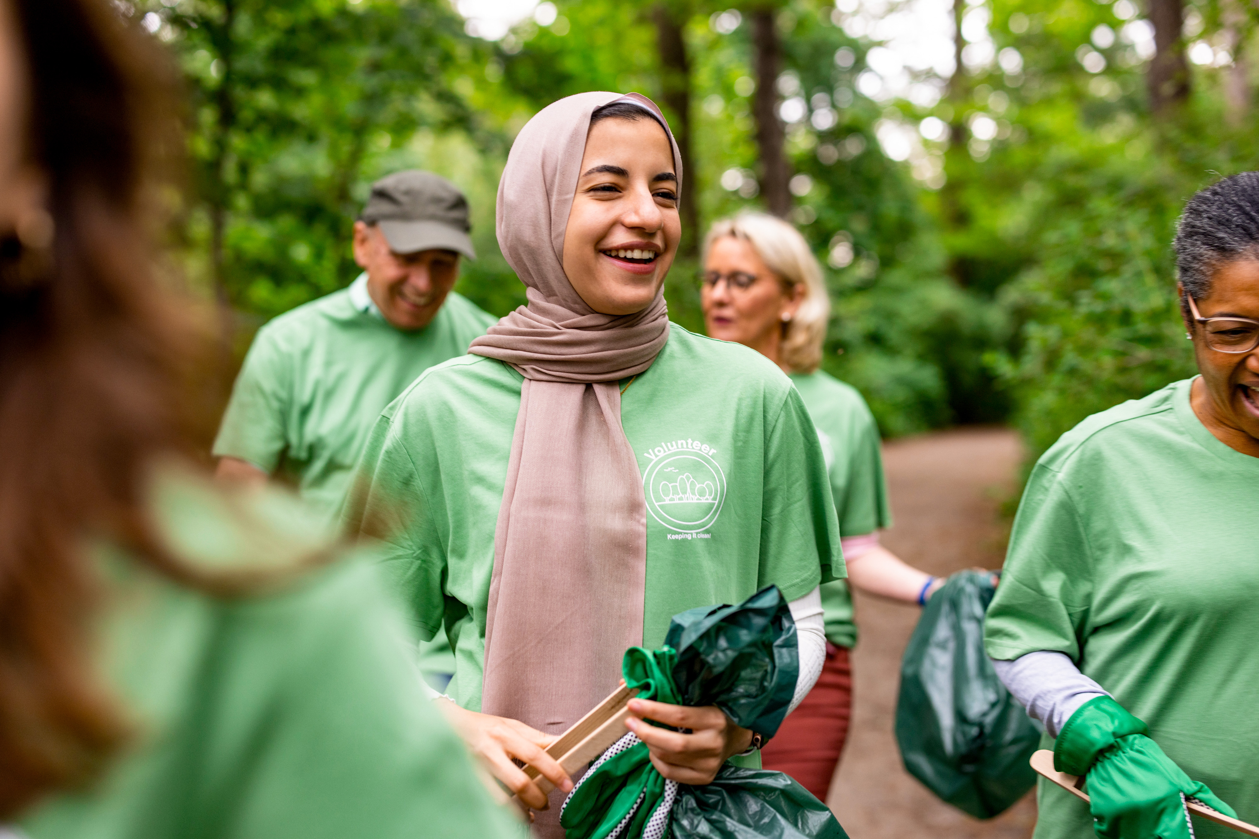 A group of volunteers wearing green shirts and holding trash bags are cleaning up a forested area. A group of volunteers wearing green shirts and holding trash bags are cleaning up a forested area.