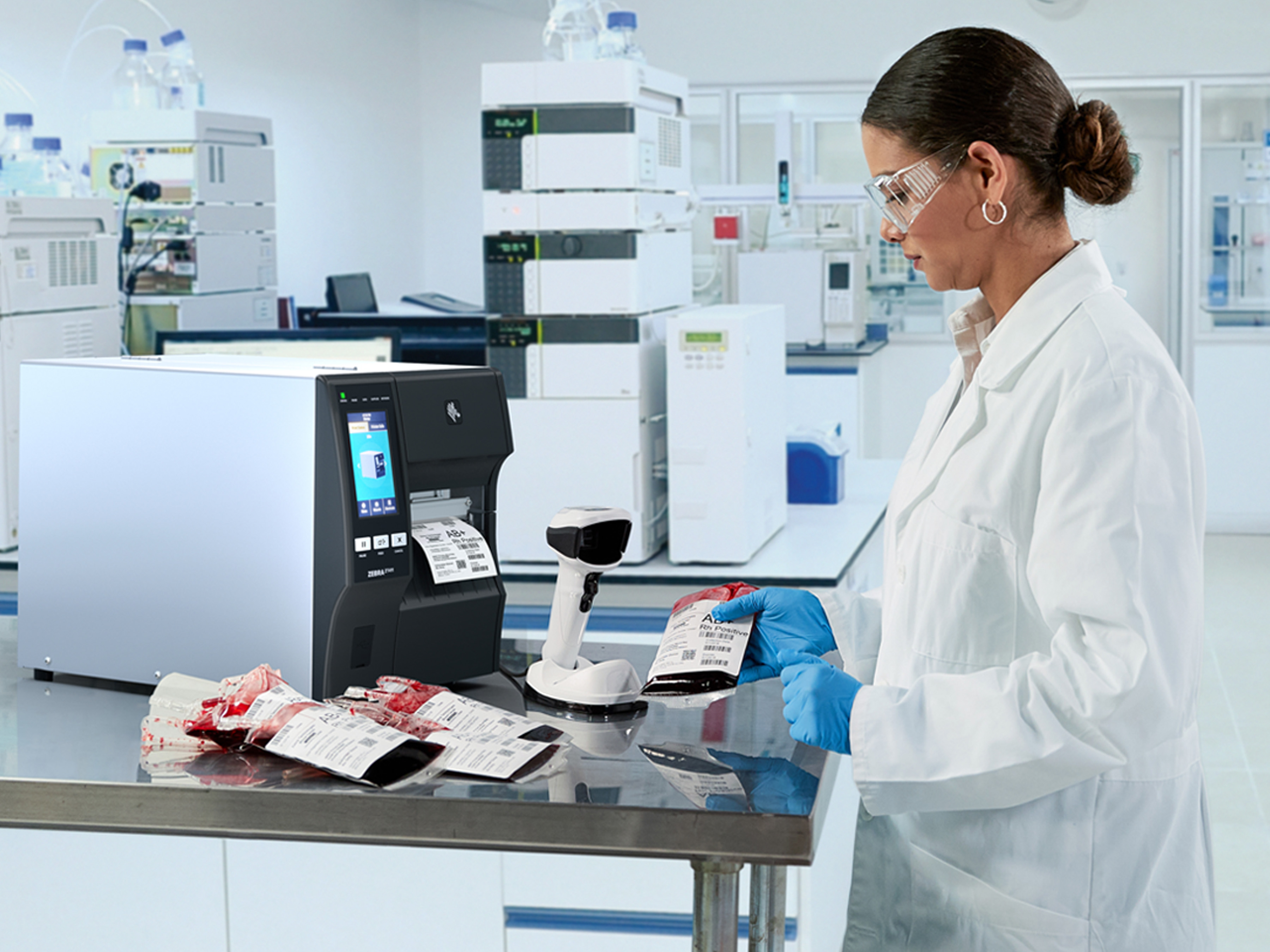 A healthcare professional is in a laboratory, holding a bag of blood to check the information on the label. She is standing next to a label printer and a handheld scanner.