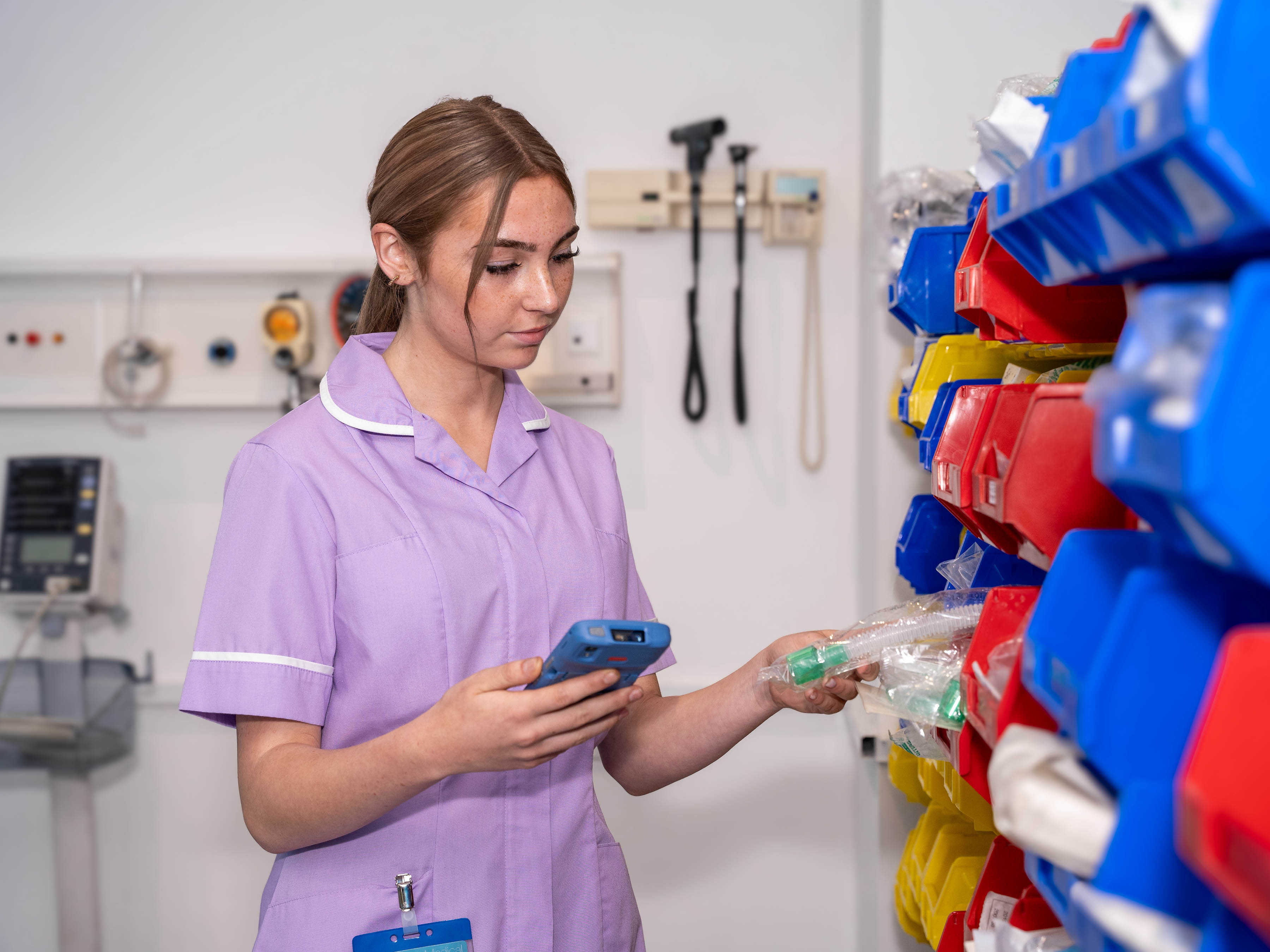 A nurse holds a clinical mobile device in her hand while looking at an item in the supplies storage area