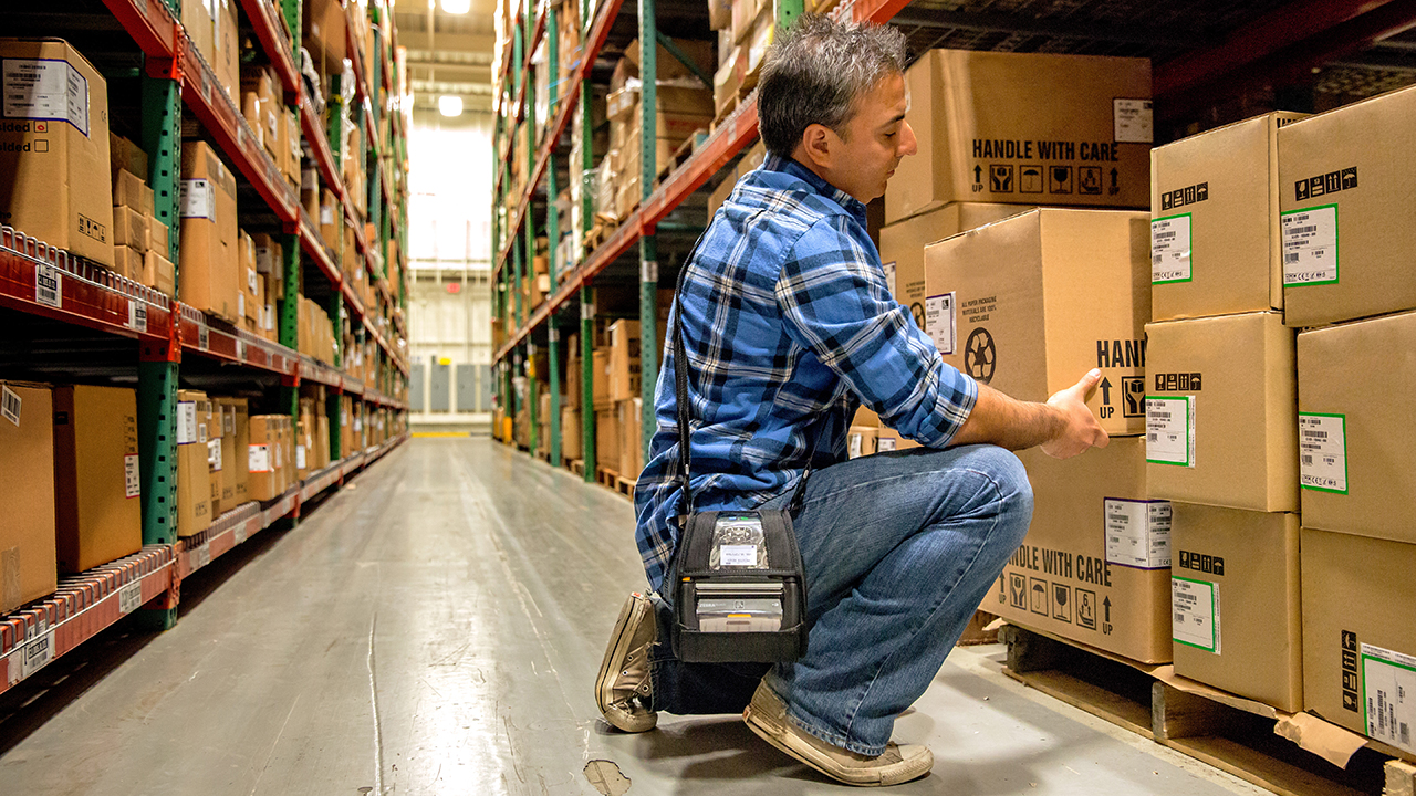 A warehouse worker carries a Zebra mobile printer on his shoulder while moving boxes