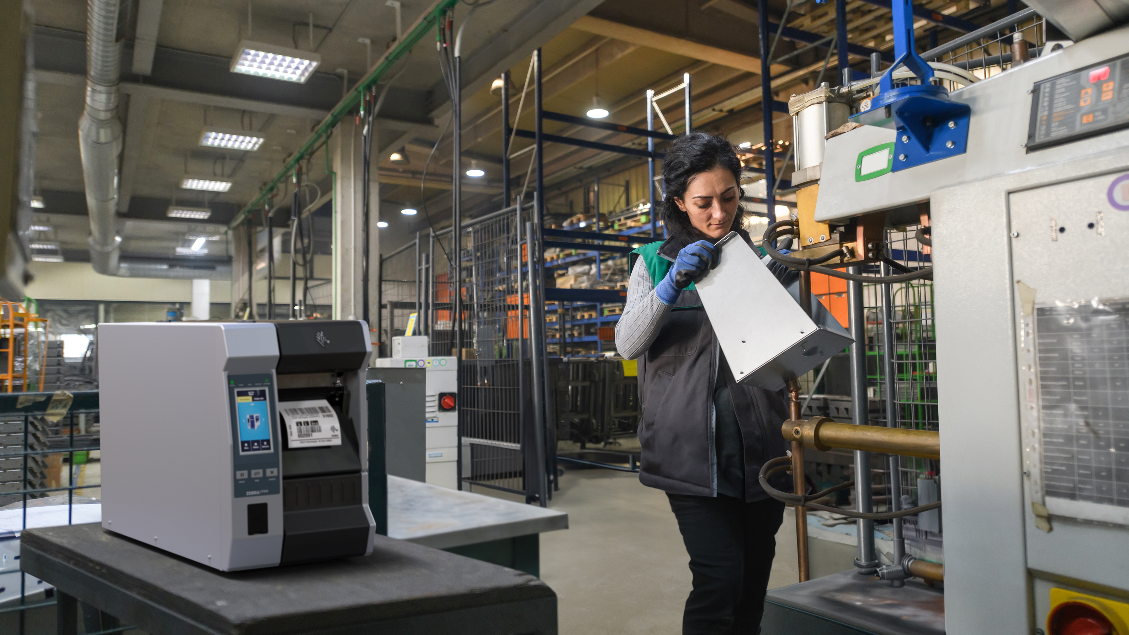 Worker in a manufacturing setting while a Zebra industrial zebra prints a label. Worker in a manufacturing setting while a Zebra industrial printer prints a label.