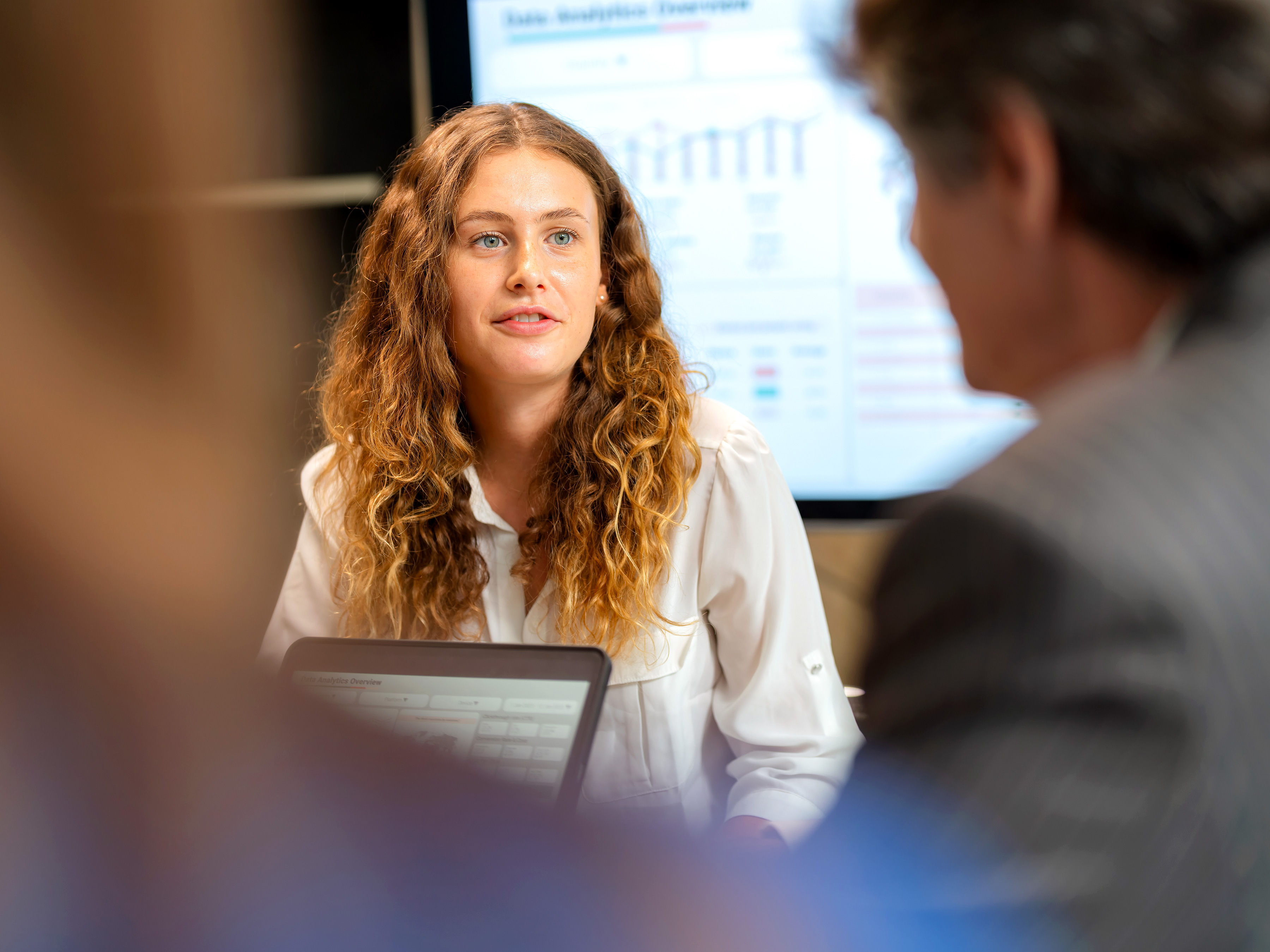 Woman leading a meeting with a laptop on hands and a chart on screen in the background Woman leading a meeting with a laptop on hands and a chart on screen in the background