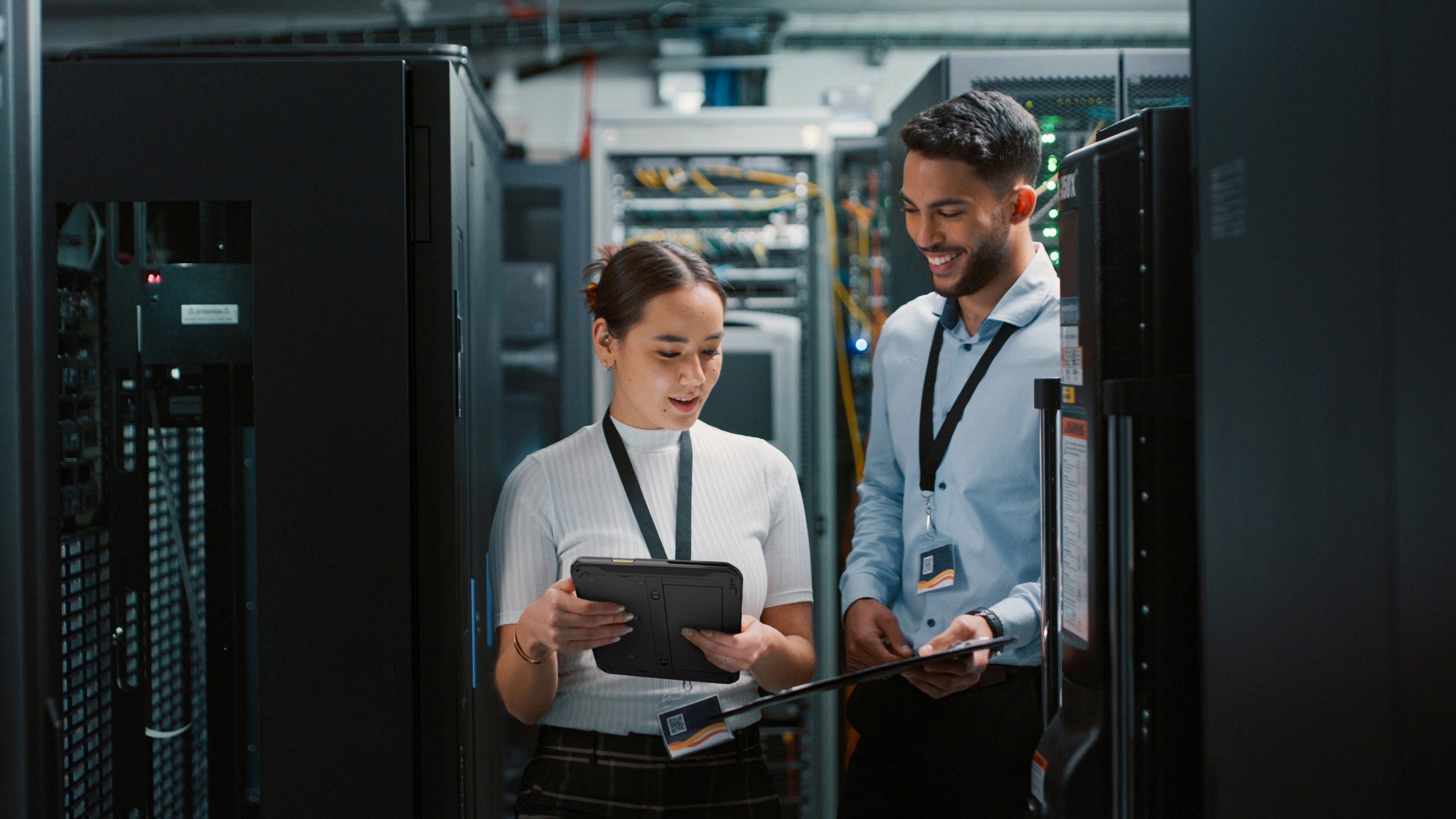 Two coworkers reviewing their Zebra tablet and handheld mobile computer inside of a server room. Two coworkers reviewing their Zebra tablet and handheld mobile computer inside of a server room.