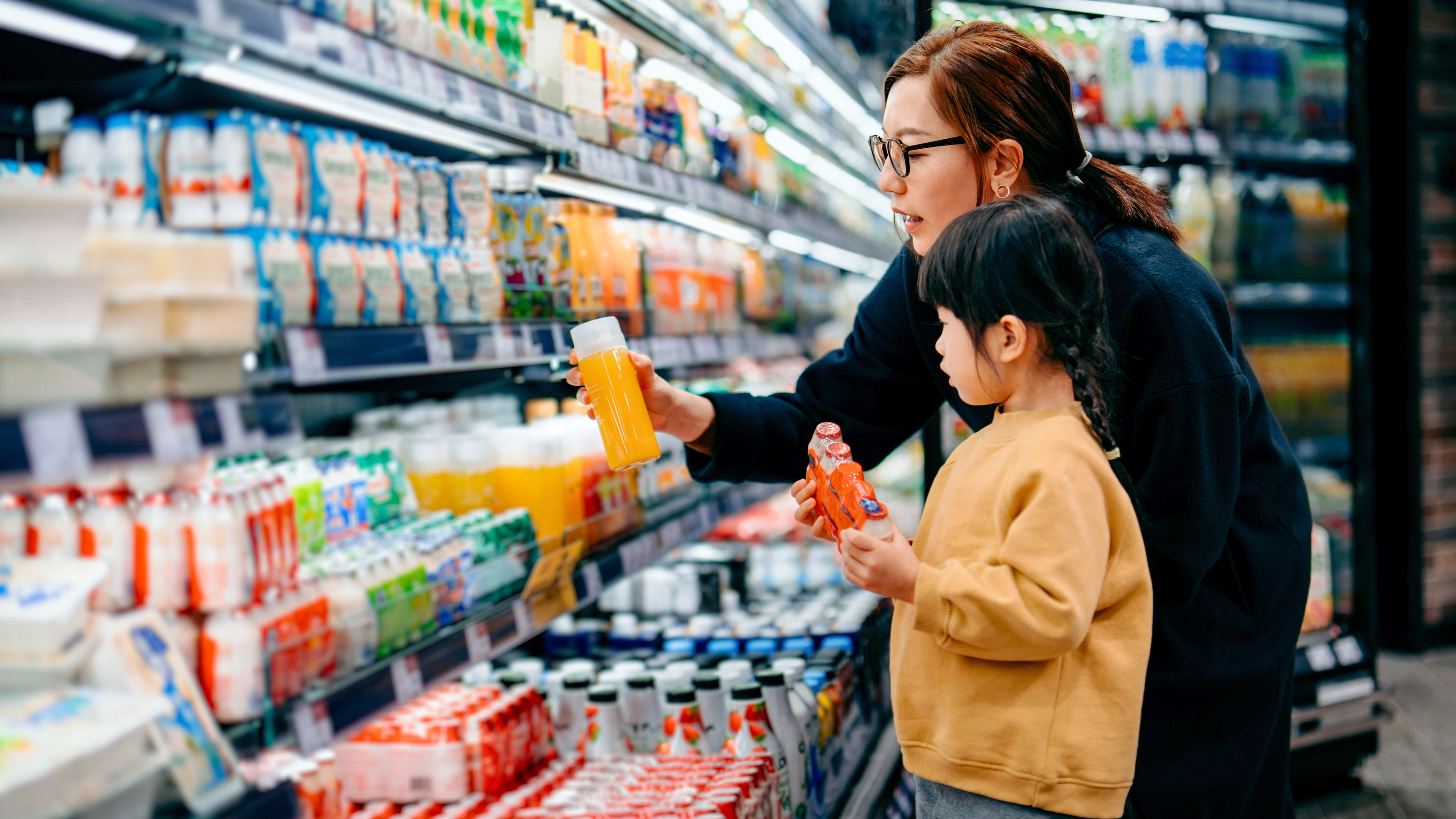 A parent and a child are shopping for beverages in front of a refrigerated section in a grocery store. A parent and a child are shopping for beverages in front of a refrigerated section in a grocery store.