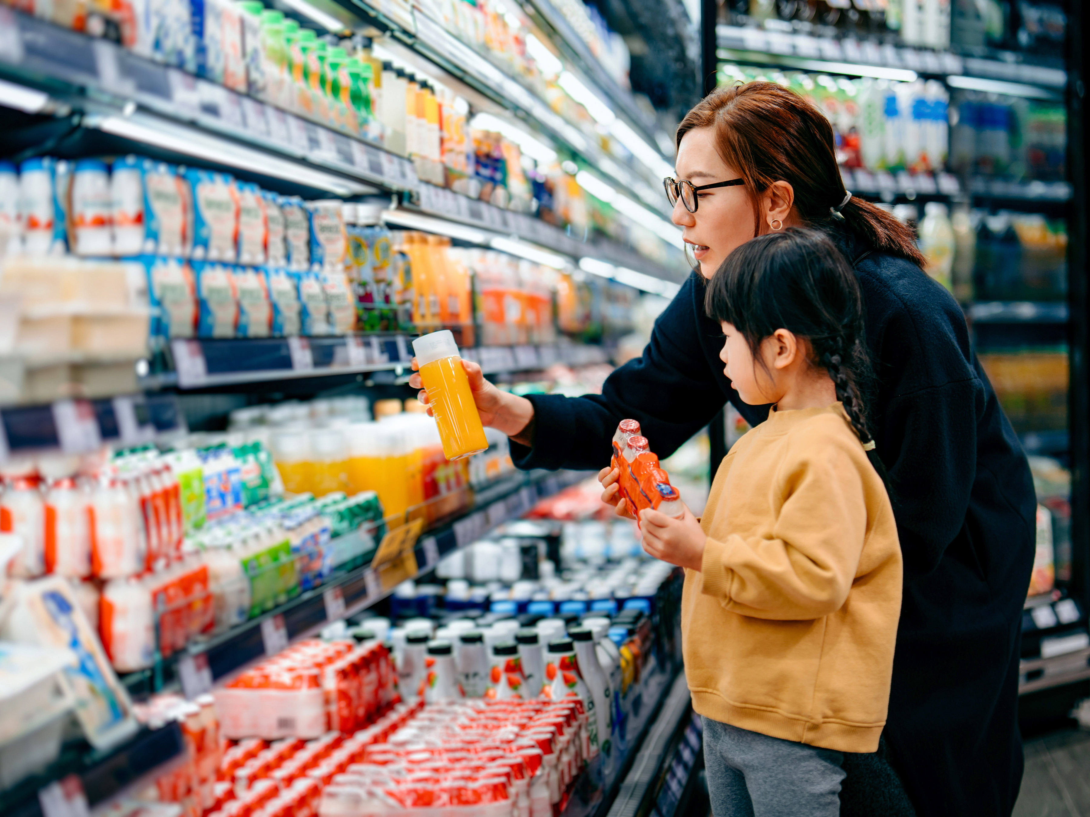 A parent and a child are shopping for beverages in front of a refrigerated section in a grocery store. A parent and a child are shopping for beverages in front of a refrigerated section in a grocery store.