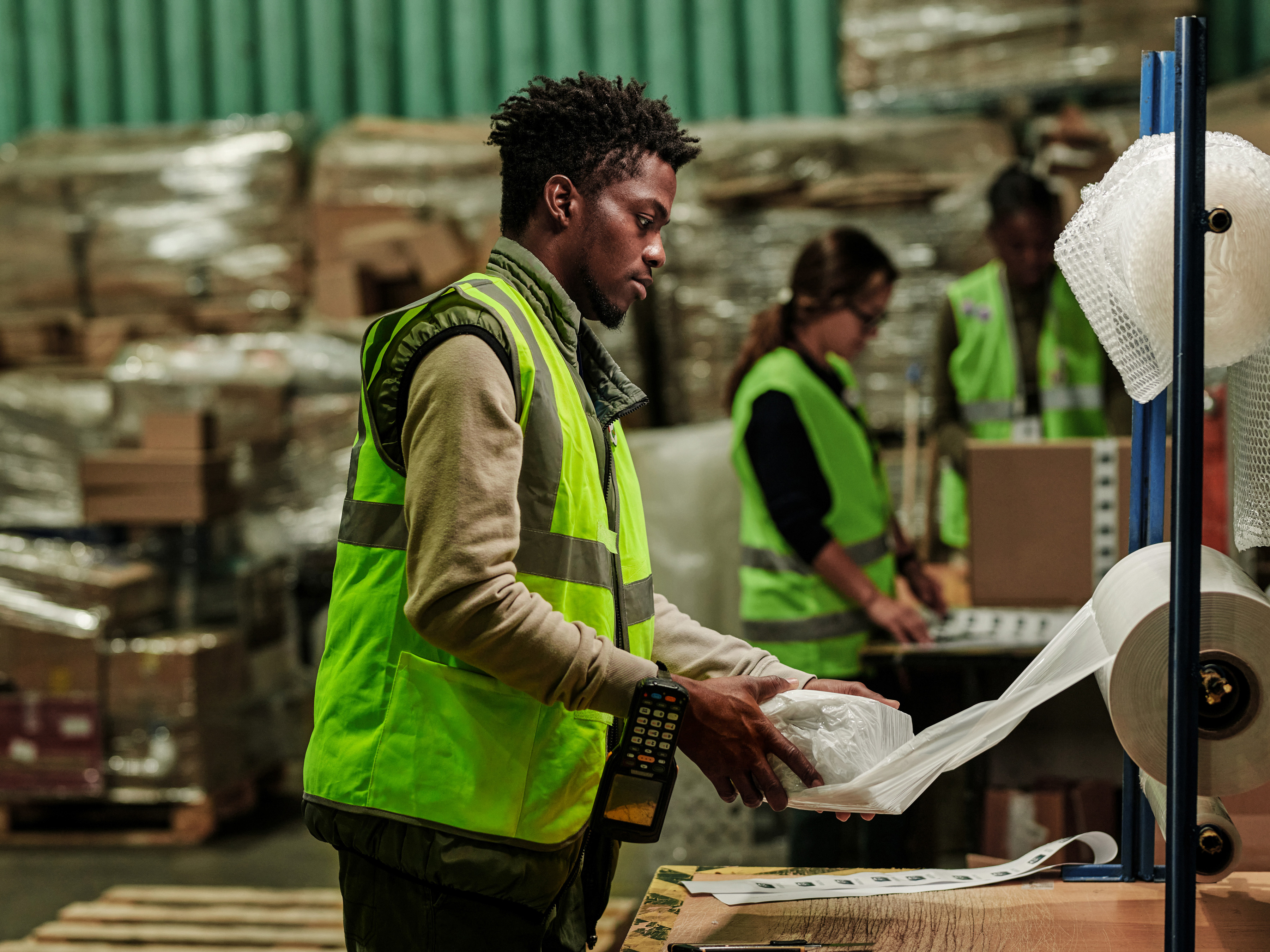 A worker in a warehouse has a handheld computer securely hanging from his wrist while packaging items using protective wrap for shipment. A worker in a warehouse has a handheld computer securely hanging from his wrist while packaging items using protective wrap for shipment.