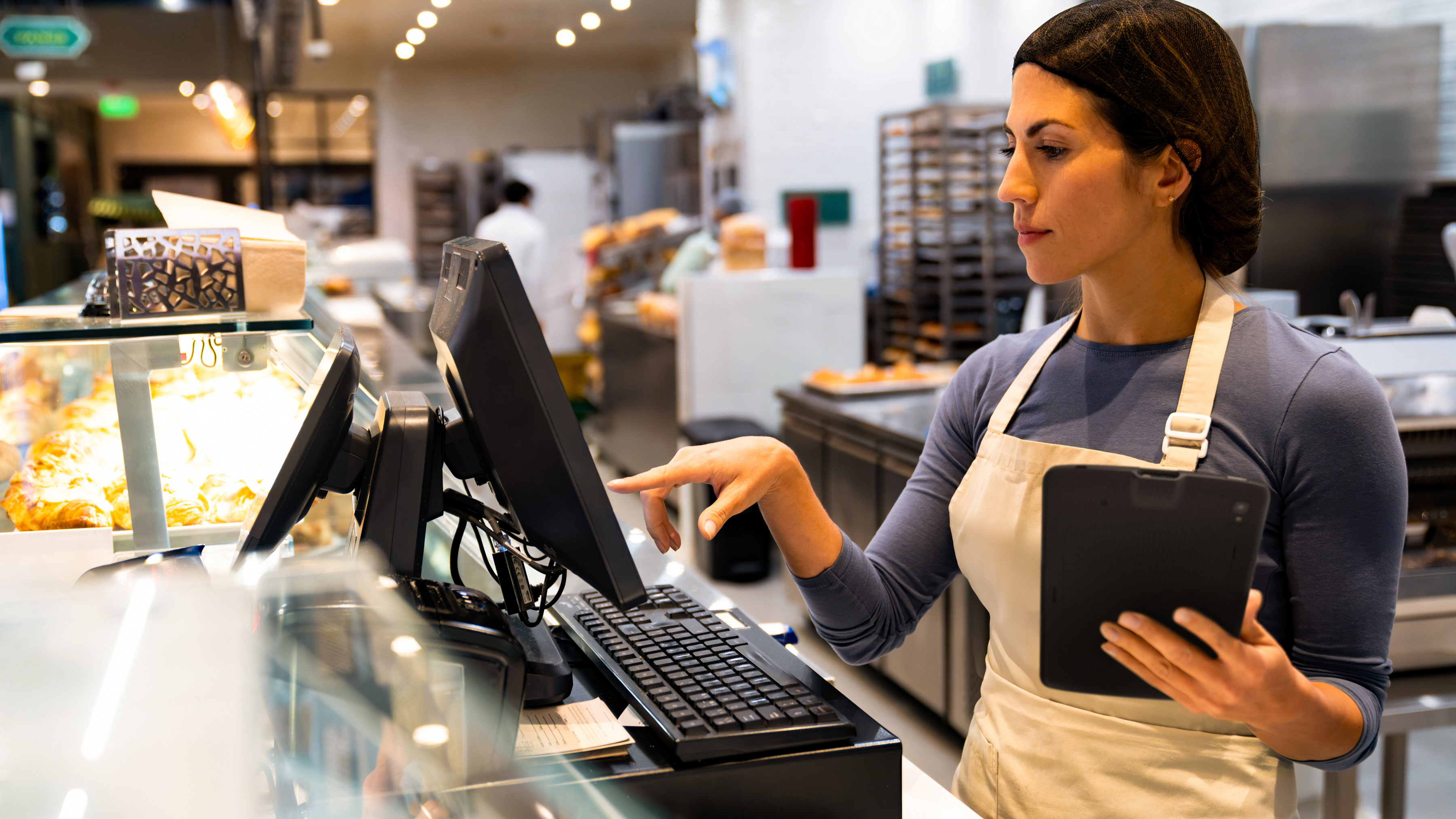 A bakery employee holding a Zebra tablet and using an interactive kiosk  for checkout at the cash register. A bakery employee holding a Zebra tablet and using an interactive kiosk  for checkout at the cash register.
