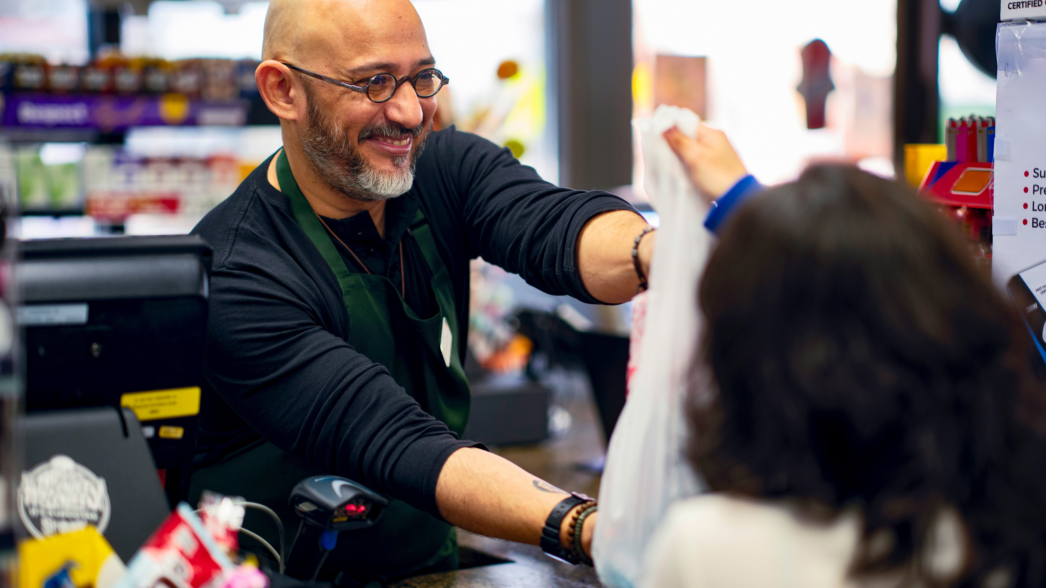 A cashier hands a plastic bag containing products to a customer at a checkout counter in a convenience store. A cashier hands a plastic bag containing products to a customer at a checkout counter in a convenience store.
