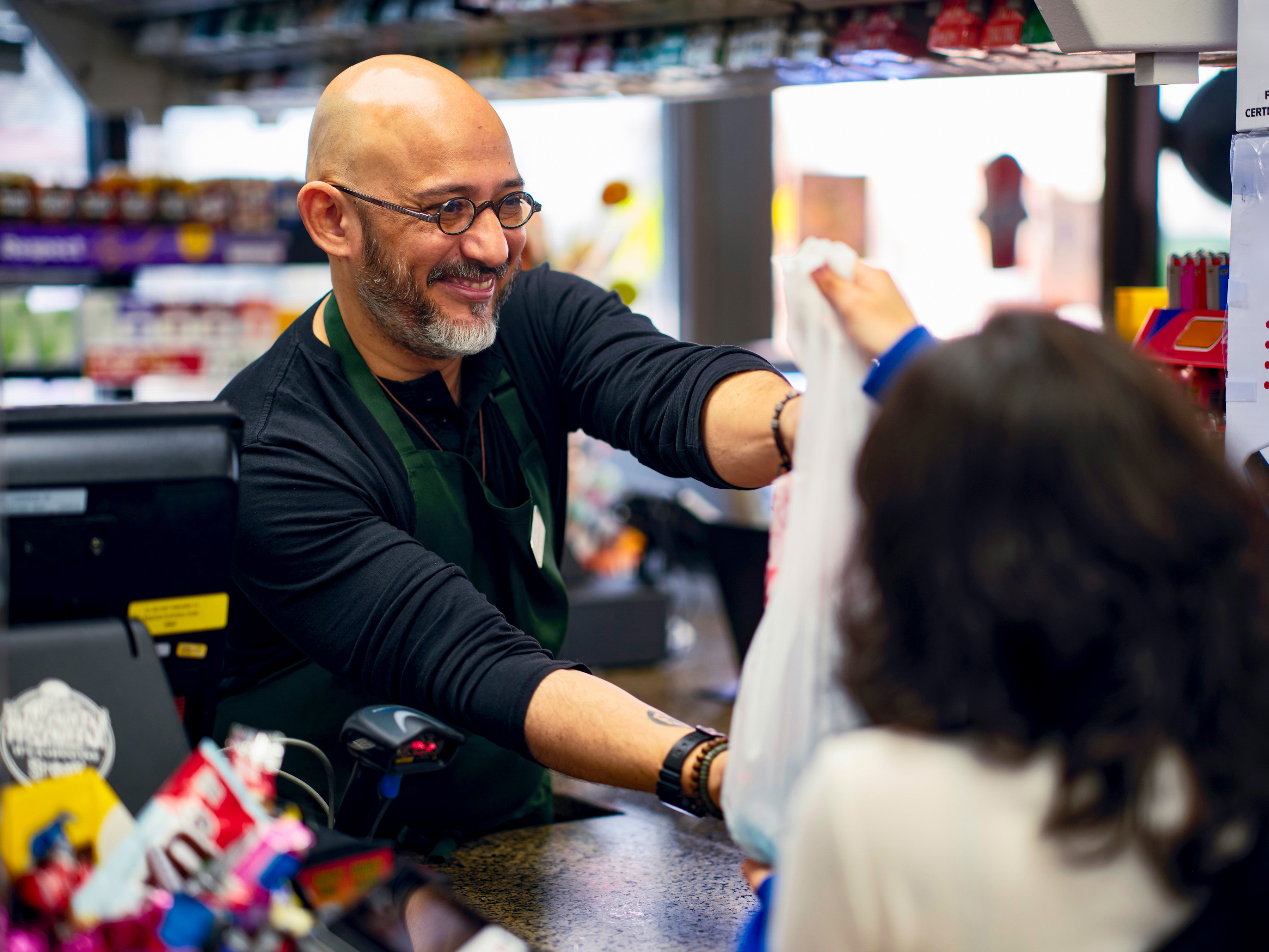 A cashier hands a plastic bag containing products to a customer at a checkout counter in a convenience store. A cashier hands a plastic bag containing products to a customer at a checkout counter in a convenience store.