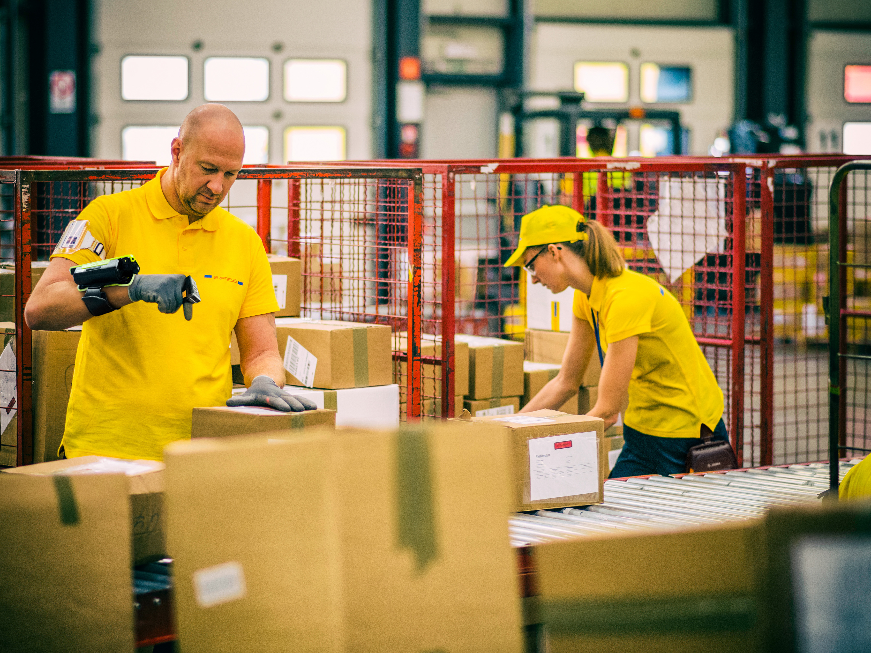 Two postal workers in yellow uniforms are sorting and scanning boxes using wearable scanners. Two postal workers in yellow uniforms are sorting and scanning boxes using wearable scanners.