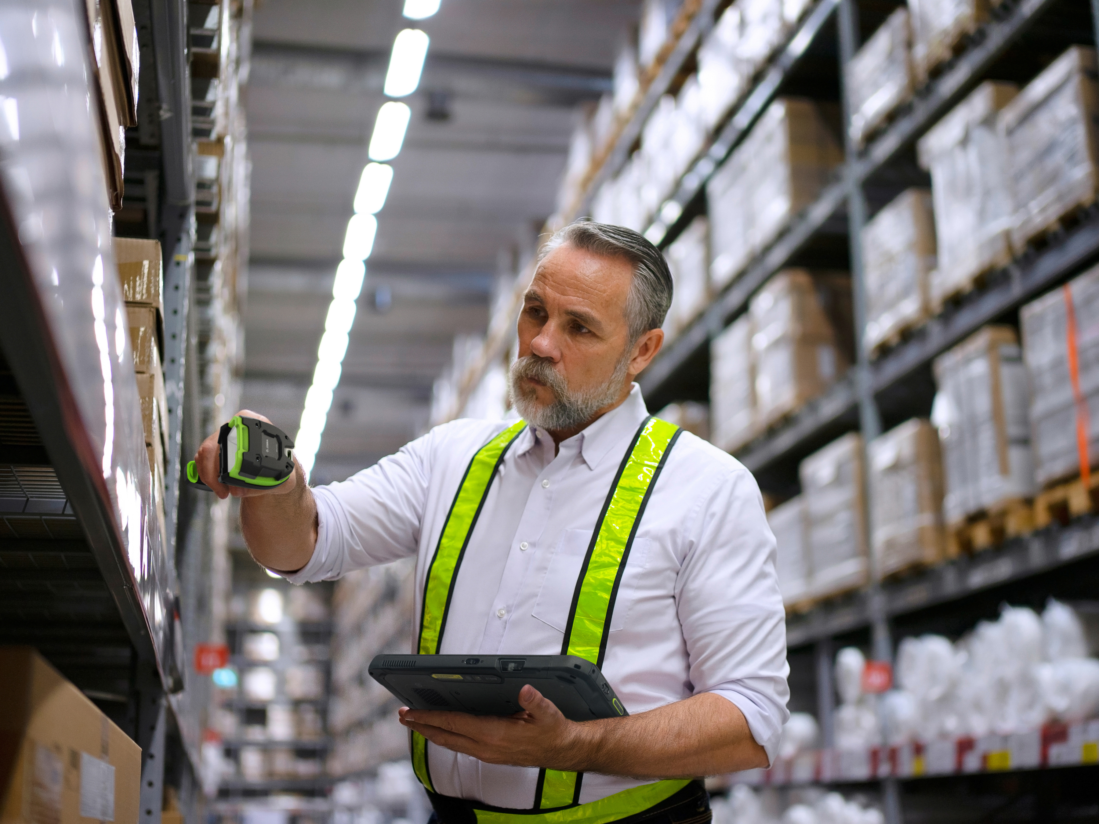 An employee using a handheld barcode scanner and a tablet to scan shelf stock inside of a warehouse. An employee using a handheld barcode scanner and a tablet to scan shelf stock inside of a warehouse.